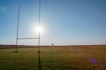 Fototapeta na wymiar Field with tall goal posts for Irish National sports on green grass against clear blue sky and the sun. Rugby, hurling, camogie and Gaelic football training ground. Nobody. Popular sport in Ireland.