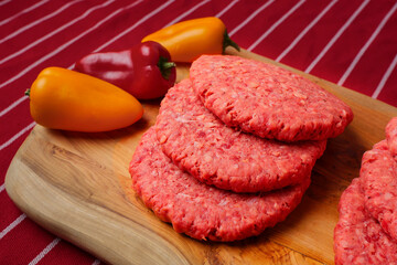 Stack of fresh uncooked beef burgers on a wooden cutting board and red and yellow peppers on red and white stripe butcher apron. Meat industry product for mass consumption. - obrazy, fototapety, plakaty