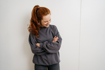young redhead woman stands laughing with crossed arms in front of a white wall and looks down