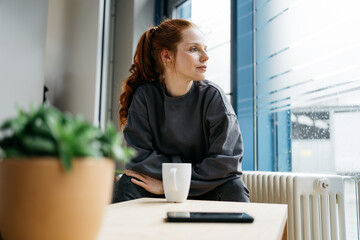 young redhead woman takes break in her office and looks thoughtfully to the side - 525763427