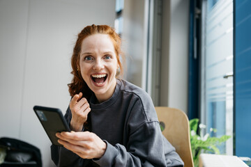 Fototapeta premium young redhead woman with cell phone looks at camera in amazement