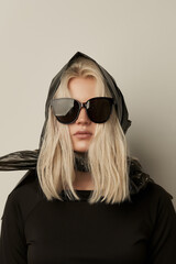 Naklejka premium Close-up shot of a girl wearing black sunglasses with wide temple. The blonde girl in a kerchief, black sunglasses and a black top are on a pastel ivory background. Front view.