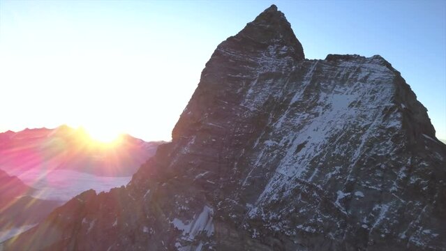 Drone view of peak of the Matterhorn in the Alps. Mont Cervin at sunrise. Aerial view flying toward jagged mountaintop with snow on cliff face and rising sun background.