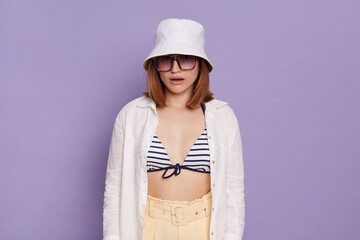 Portrait of confused surprised woman wearing panama, white shirt and swimming suit, looking at camera with dropped jaw, posing isolated over purple background.