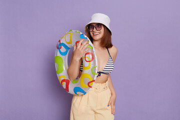 Fototapeta premium Portrait of pretty satisfied woman with rubber ring wearing panama and swimming suit posing isolated over purple background, looking away with toothy smile, enjoying her vacation.