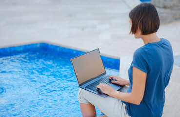 Young business woman working at computer by hotel pool. Young lady downshifter working at laptop and enjoys and relaxed environment, working day. Online freelance work on vacation.