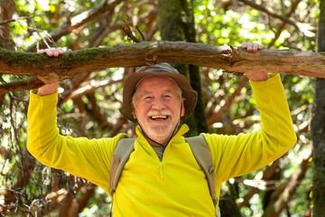 Cheerful elderly man with hat and backpack hanging from a tree branch looking at the camera smiling while hiking in the woods
