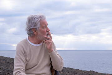Black and white portrait of senior man sitting on the cliff in sea excursion smoking a cigarette...