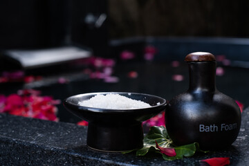 Bath salt in the bowl and bath foam in ceramic bottle on bathtub full of rose petals. Spa concept. Skin care treatment in spa salon. Wellness, healthy lifestyle, relaxation procedure. Selected focus.