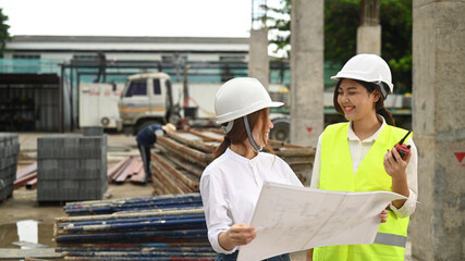 Smiling female real estate investor and civil engineers manager wearing safety helmets examining plans at construction site