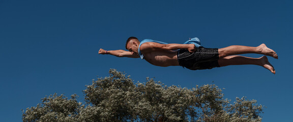 Young teen boy wearing a towel as a superhero scarf flying and diving in river. Clear blue sky and...
