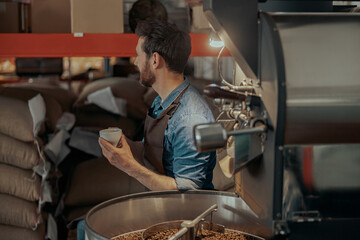 Business owner with cup of fresh coffee on background of roasting machine looking away