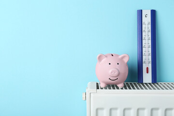 Piggy bank and thermometer on heating radiator against light blue background, space for text