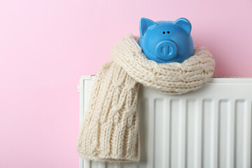 Piggy bank wrapped in scarf on heating radiator against pink background, space for text
