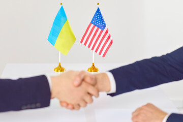 Diplomats from USA and Ukraine exchange handshake at office table with two national flags in...