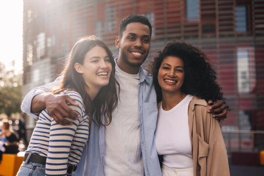 Smiling young caucasian and african friends hugging and posing standing outdoors. Brunettes guy and girls wear casual clothes. Relaxed lifestyle, concept