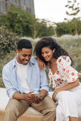 Nice young african friends listen to music in headphones, look at phone sitting on street. Brunette guy wears shirt, girl wears blouse. Concept of use, leisure