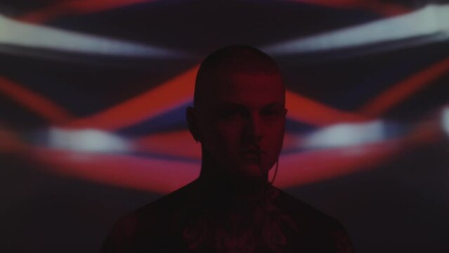 Chest up shot of tattooed man with ear to nose chain dancing to music in dark studio with blinking neon light and kaleidoscopic projection in background