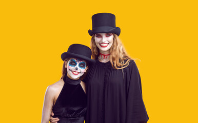 Happy family with Halloween creative makeup. Joyful young woman and her teenage daughter celebrating traditional Halloween holiday together. Family in festive spooky outfits on orange background.