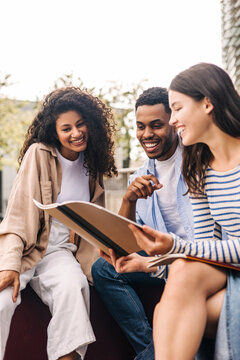 Group of young interracial students discussing homework looking at exercise book sitting on campus. Brunette guy and girls wear spring casual clothes. Education concept