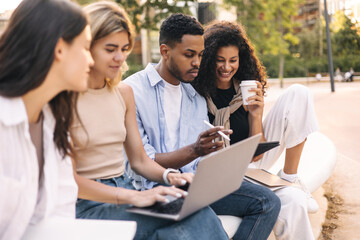 Attentive young intercultural guy with girl doing analysis of work done using laptop. Students study after class sitting outdoors. Education concept