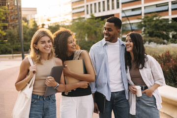 Cheerful young friends of caucasian, african appearance are walking around sunny city after class. Boy and girls carry educational things with them. Teenage concept