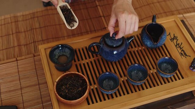 Traditional utensils laid out on a wooden tray in preparation of tea ceremony. Hands are picking up scoop full of tea leaves and a wooden stick and pour dry tea leaves into a teapot. Shot from above