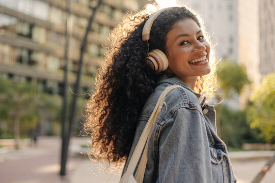 Young beautiful african woman listening to songs in headphones walking outdoors. Brunette with curly hair wears denim jacket. Enjoying, music concept.