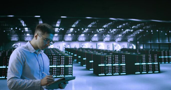 A young man wearing glasses using tablet while working in server room in contemporary data center.