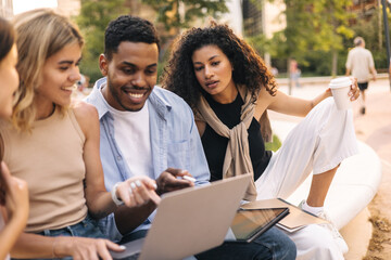 Interested young interracial people looking at gadget screen working on laptop outdoors. Students wear casual clothes spending time with friends. Leisure concept