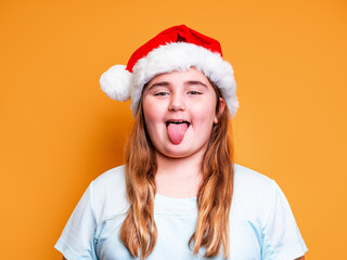 adorable little girl sticks out her tongue dressed up in a santa claus hat on a yellow background