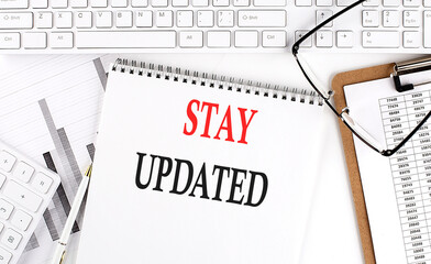 Text STAY UPDATED on Office desk table with keyboard, notepad and analysis chart on white background.