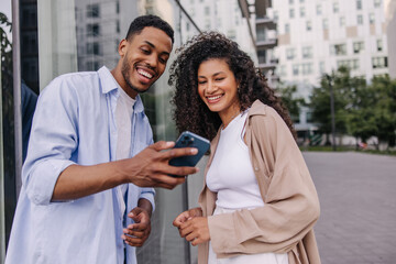 Two young african students are using modern smartphone relaxing outdoors. Brunette guy and girl...