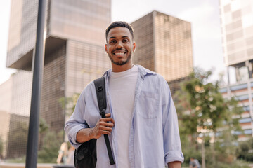 Smiling young african guy smiling looking at camera relaxing outdoors. Brunette man with stubble wears blue shirt. City life concept