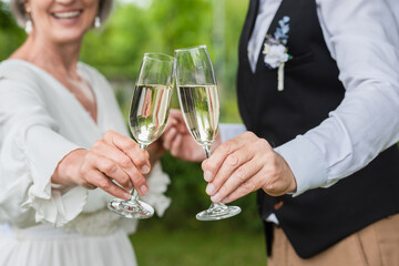 cropped view of happy and mature newlyweds clinking glasses of champagne in green garden