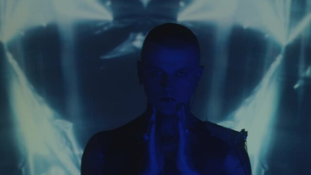 Chest up shot of tattooed man with ear to nose chain dancing in dark studio with blinking blue neon light and kaleidoscopic projection in background