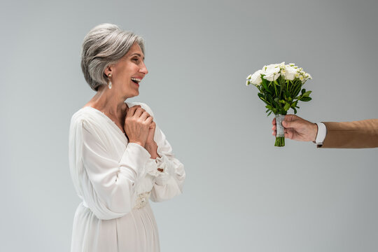 groom holding wedding bouquet near happy middle aged bride in white dress isolated on grey