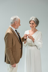 cheerful mature bride in white dress wearing golden engagement ring on finger of middle aged groom isolated on grey
