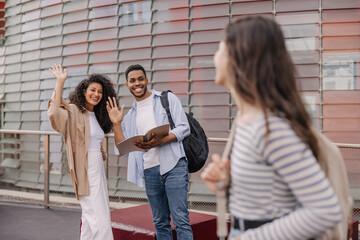 Positive young african guy and girl greet girlfriend spending time outdoors. Brunette students wear casual clothes in spring. Real emotions concept