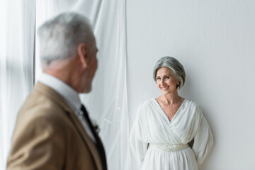 blurred man in suit looking at cheerful mature bride in white dress near white curtains
