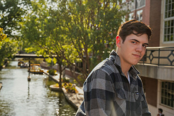 Handsome young man dressed in a plaid jacket posing in front of a river walk.