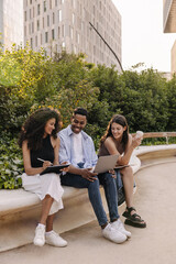 Happy young interracial students use laptop and tablet preparing for their lessons outdoors. Brunette guy and girls wear casual clothes in summer. Technology concept