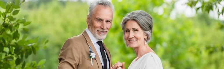 happy middle aged groom in suit holding hands with mature bride in garden, banner - Powered by Adobe