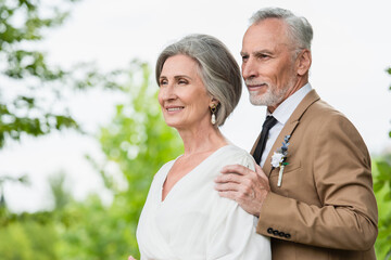 pleased middle aged groom in suit hugging mature bride in white dress in garden - Powered by Adobe