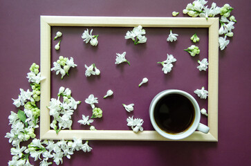 Flatlay dark purple background with small white lilac flowers? white coffee cup on textured background inside a wooden frame, diagonal corners of white lilac flowers around