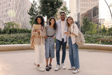 Full length positive young interracial friends posing looking at camera standing outdoors. Students wear casual light-colored clothing and backpacks. City life concept