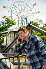 Handsome young man standing in front of iron railing and talking on a cellphone with a ferris wheel in the background.
