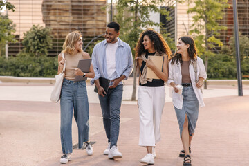 Four young intercultural guy and girls go to study, communicate with each other in open air. College students wear casual clothes spring. Lifestyle concept