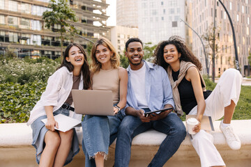 Fototapeta na wymiar Four cheerful young interracial students posing looking at camera relaxing outdoors. Guy and girls are holding tablet, laptop and notebook. Lifestyle concept