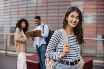 Selective focus on caucasian young girl with backpack looking at camera outdoors. Interracial university students posing near building. Lifestyle, different emotions, leisure concept